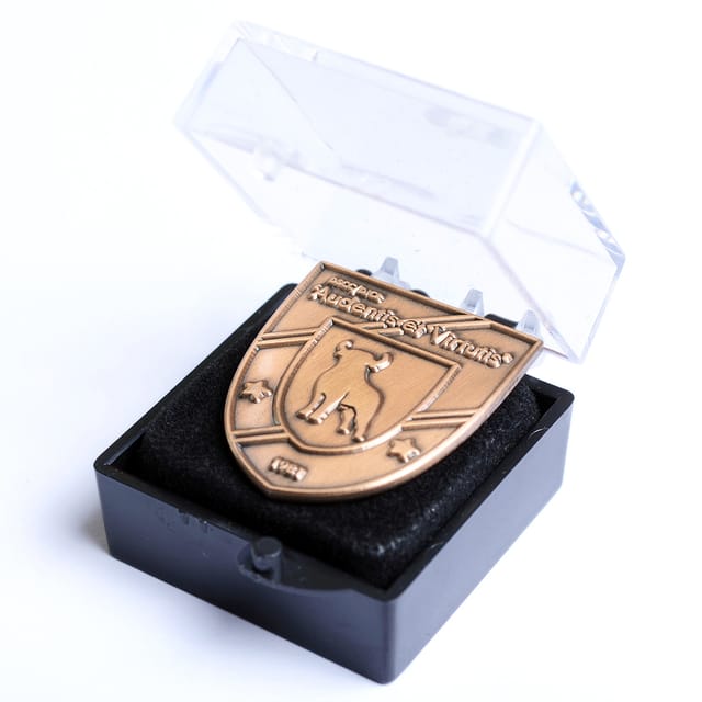 Jubileumspeldje in schildvorm in koper, in een doorzichtig geschenkdoosje